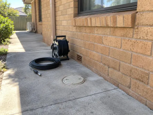 Capped drain inspection opening beside a coiled camera reel on a concrete side path by a brick house in Baulkham Hills