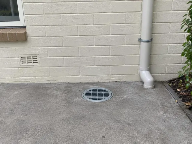Outdoor concrete gully grate beside a rendered brick wall and downpipe in Castle Hill